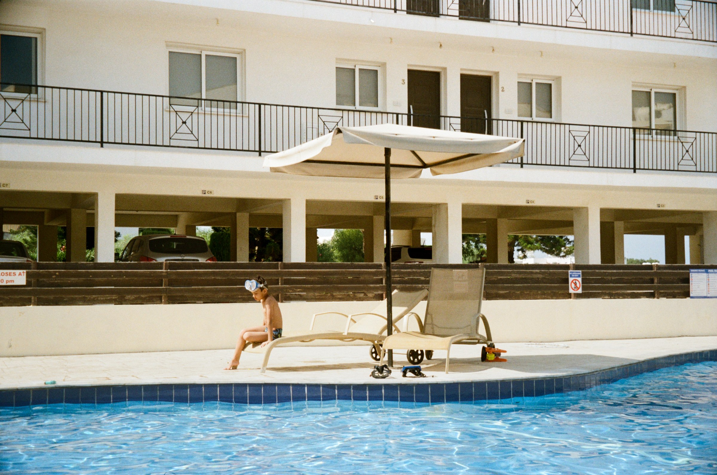 A kid sitting on a lounge chair under a poolside umbrella