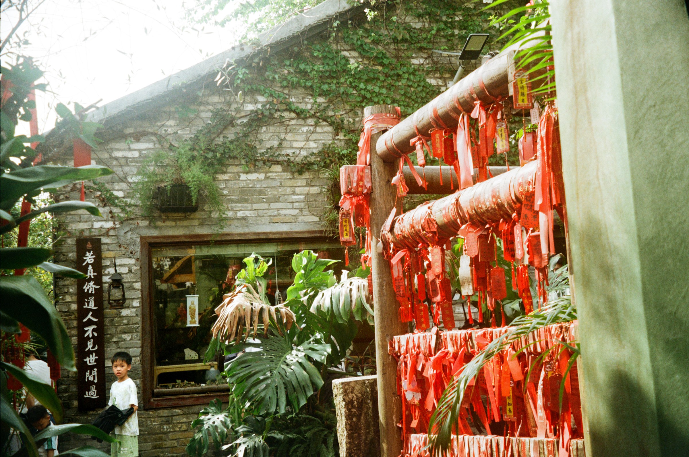 A little kid standing beside a temple wishing post hung with red ribbon plaques